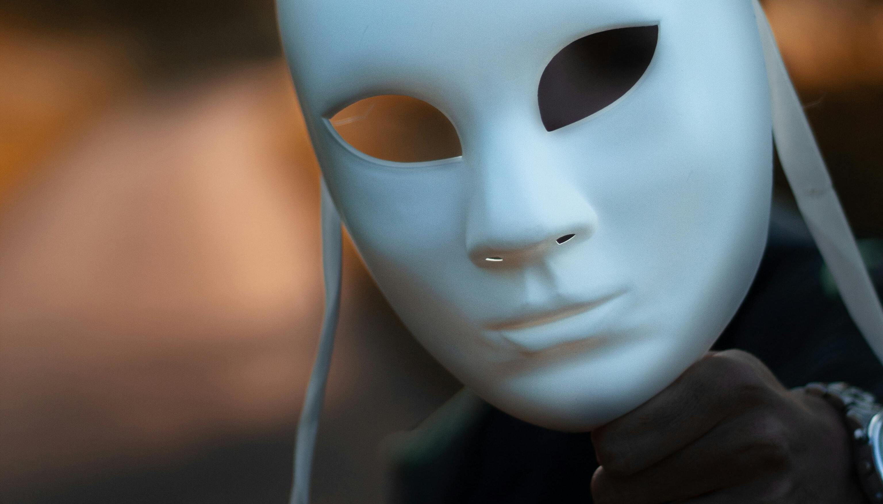 Close-up of a plain white theatrical mask held against a soft-focus warm background.